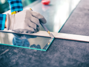 a piece of glass is laying flat on a gray surface. A person has a ruler and a cutting device and is about to make a cut on the glass