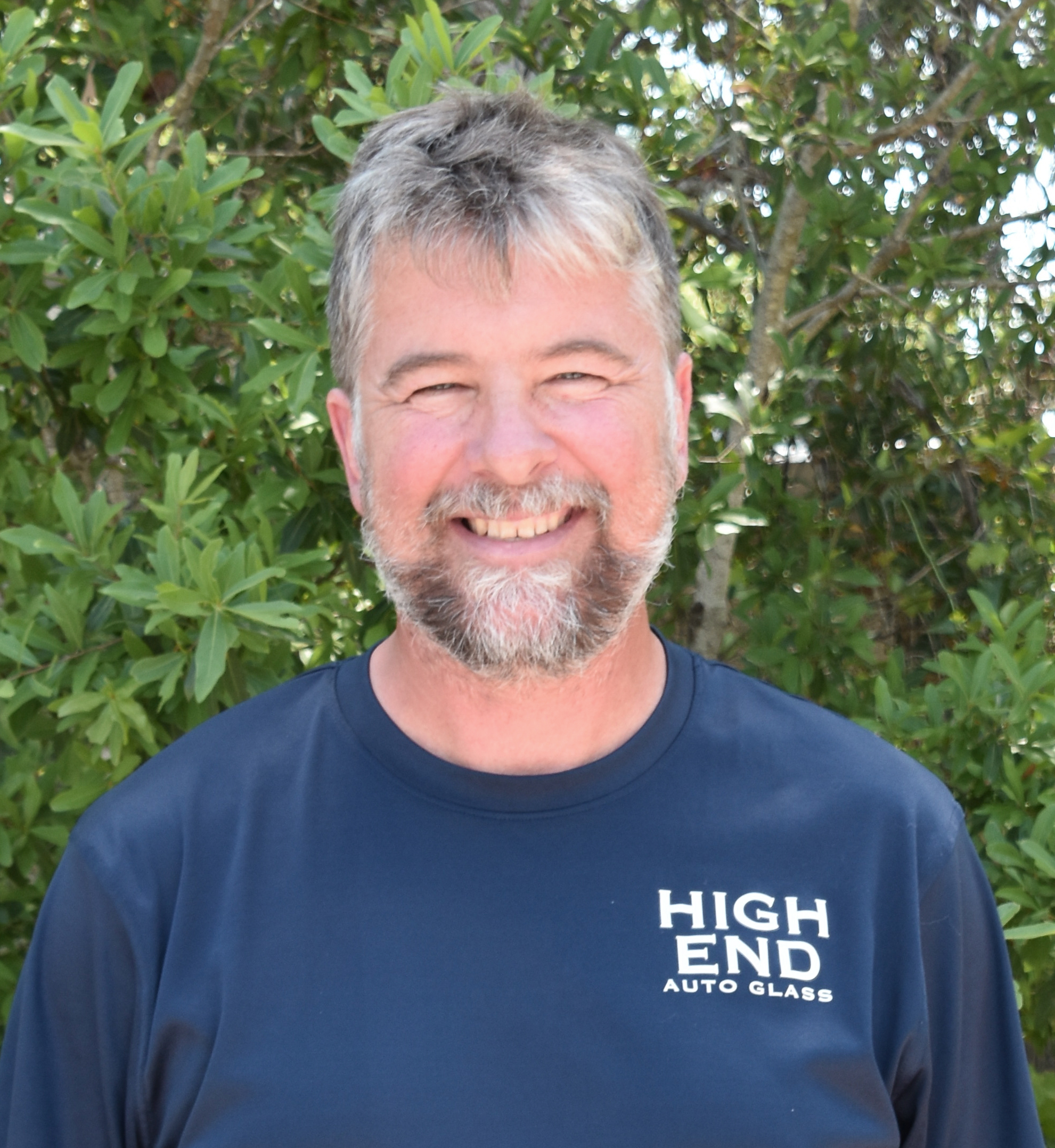 A man in a blue shirt in standing in front of a tree smiling