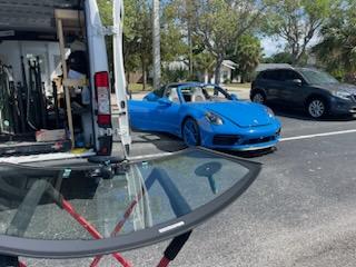A windshield is sitting on a metal stand with the open back doors of a van behind it. There is a blue sports car and a black SUV sitting to the right side of the van