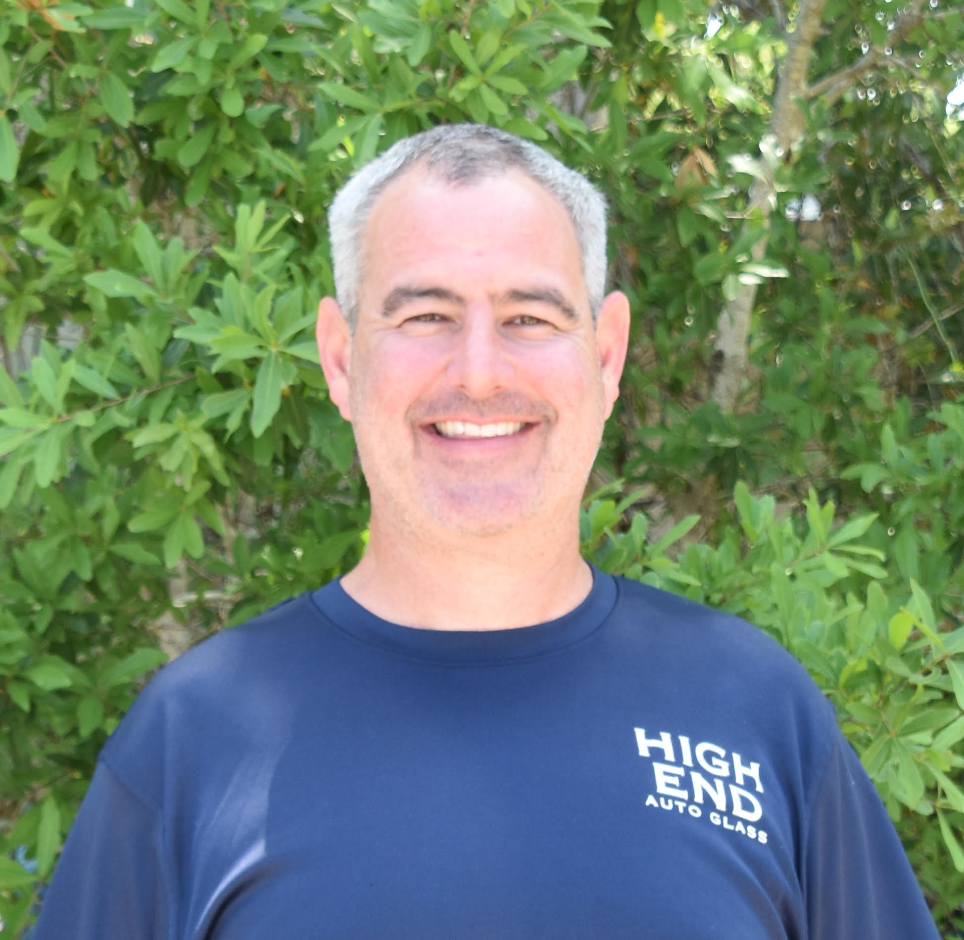 A smiling man in a blue shirt is standing in front of trees